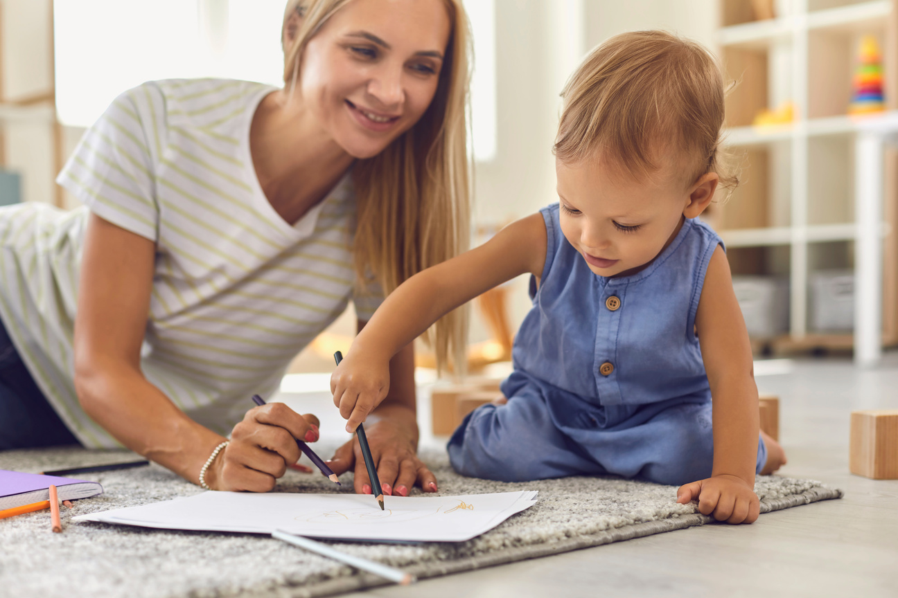 Adorable Toddler Scribbling on Paper with Pencil While Sitting on Floor with His Young Nanny